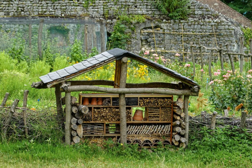 A handmade insect hotel in a lush rural garden, surrounded by vibrant flowers and rustic fencing.