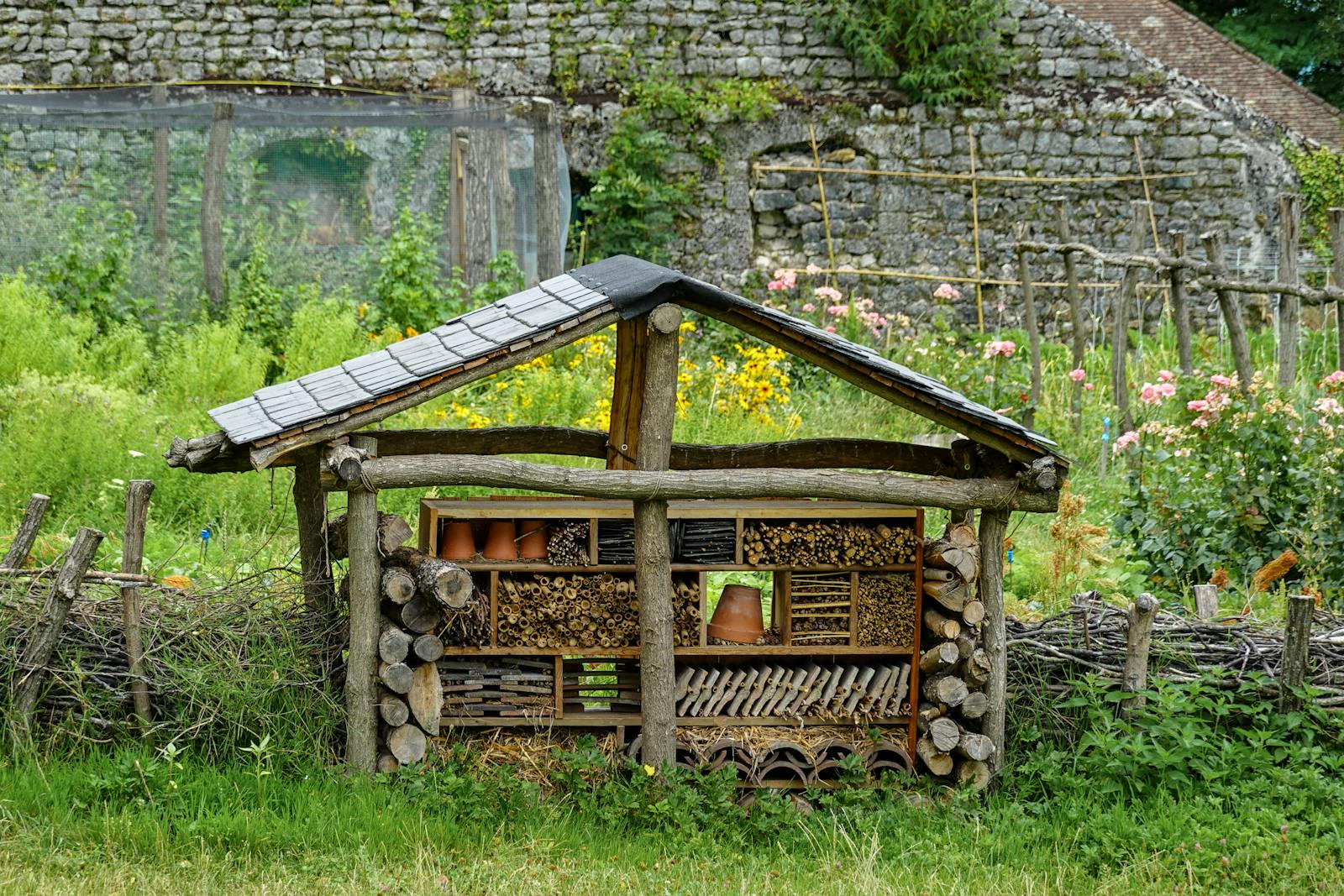 A handmade insect hotel in a lush rural garden, surrounded by vibrant flowers and rustic fencing.