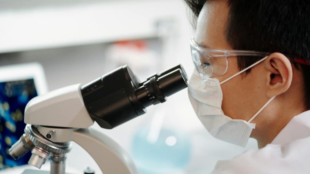 Scientist wearing face mask and goggles examining samples through a microscope in a lab.