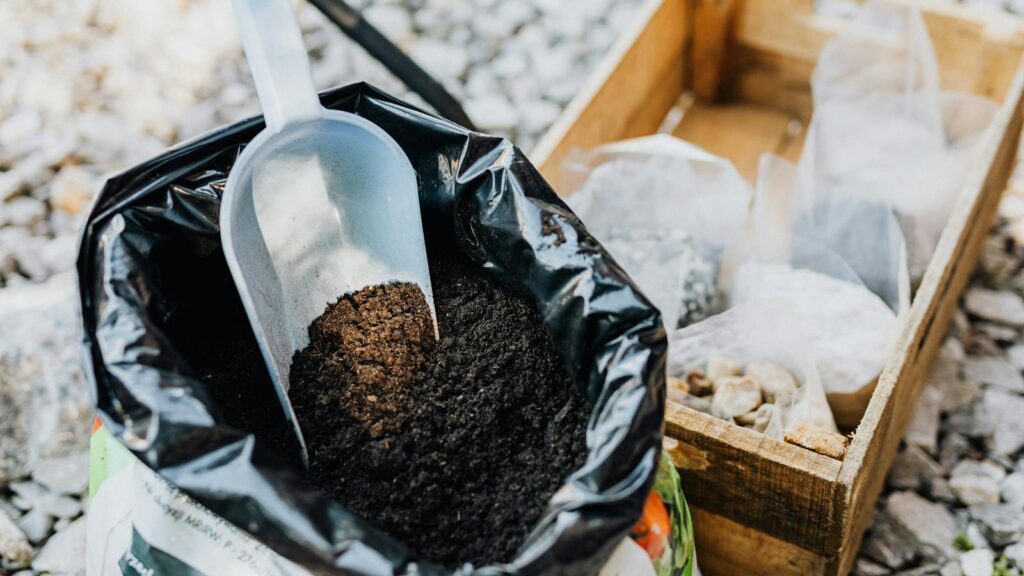 Close-up of a gardening scoop resting in a bag of rich soil beside a wooden crate filled with various gardening supplies.