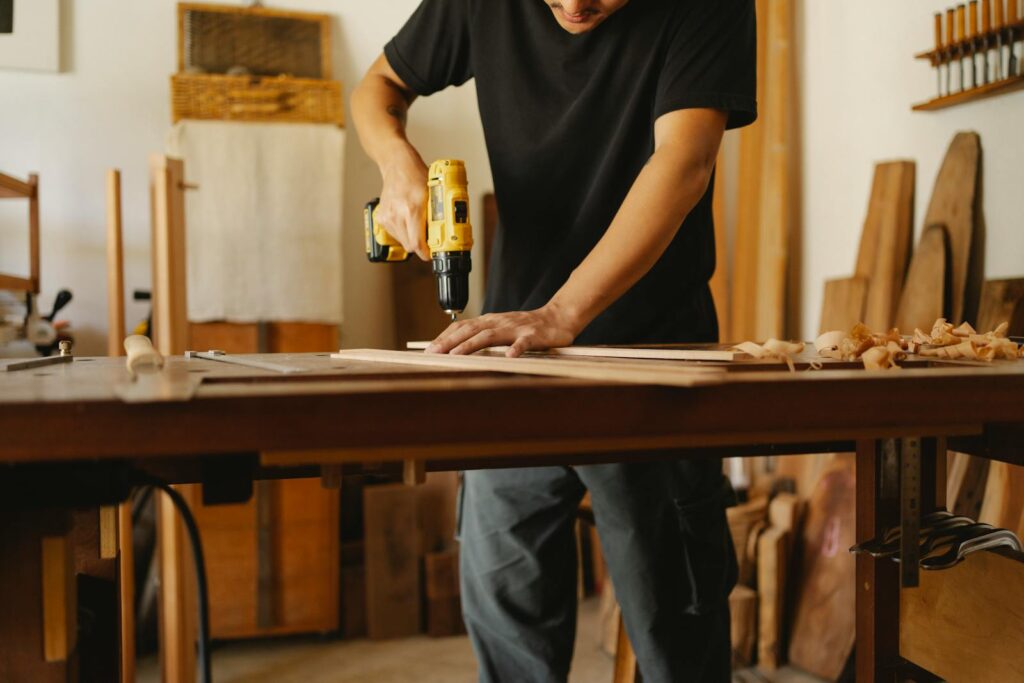 A man in a black t-shirt using a yellow cordless drill on a piece of wood in a well-lit woodworking studio.