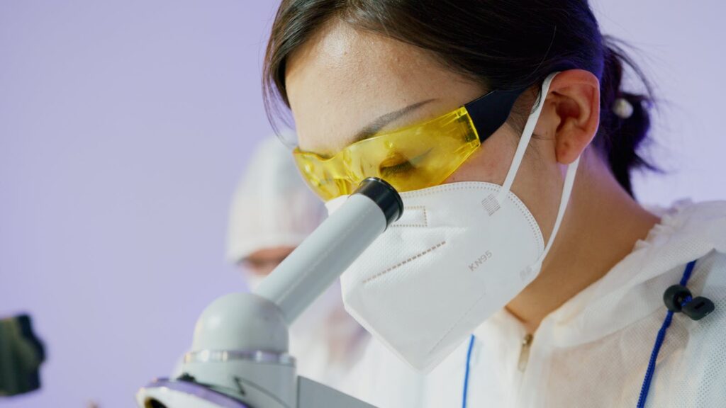 Female scientist examines specimens using a microscope, wearing protective gear.