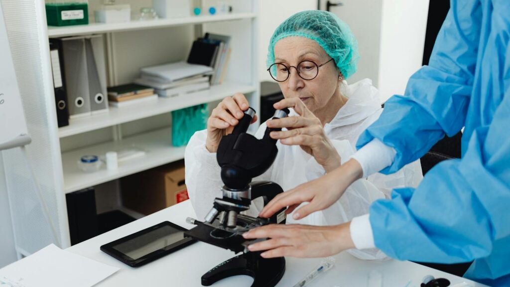 Senior researcher working with a microscope in a laboratory setting, assisted by a colleague.