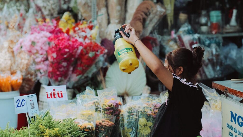 A vendor sprays yellow plant at a vibrant flower market stall. Organic floral shop scene.