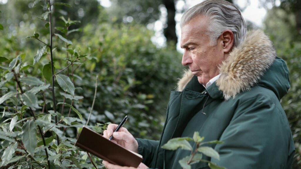 Side view of aged gray haired male in warm outerwear writing with pen in notebook while standing in park