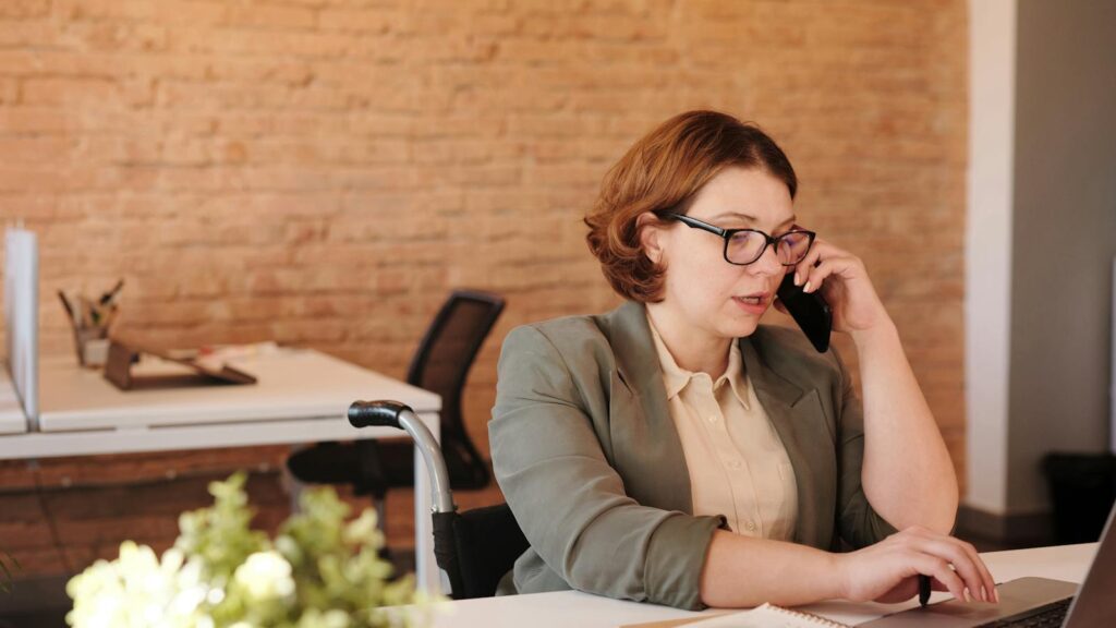 Professional woman in wheelchair working remotely on laptop and phone in a modern office.