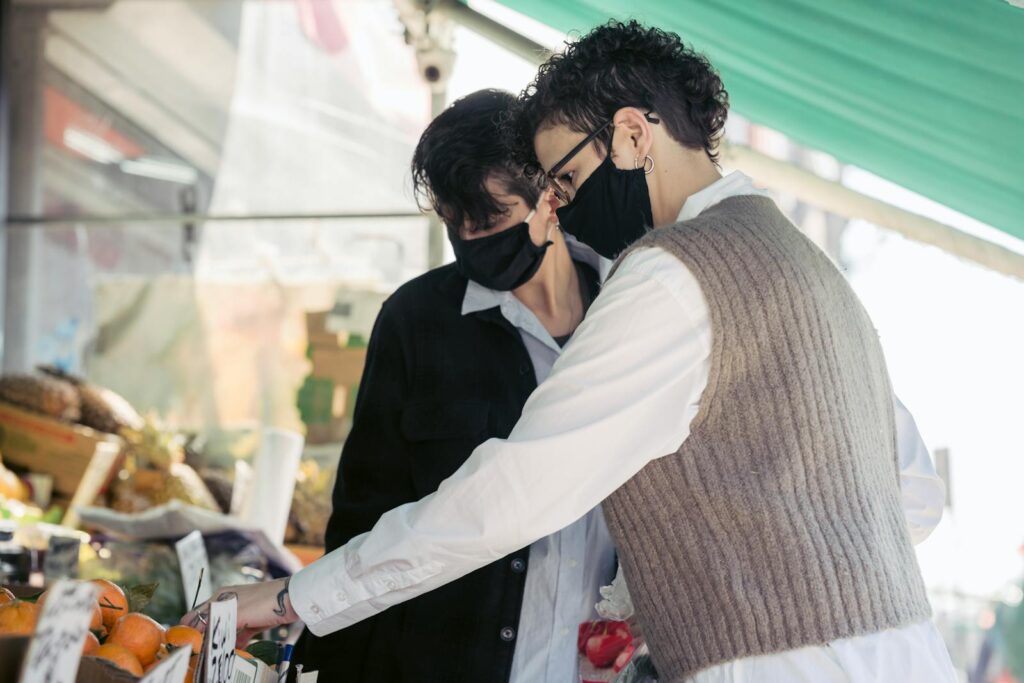 Young female friends in casual clothes and protective masks choosing fresh ripe fruits while shopping in local street market