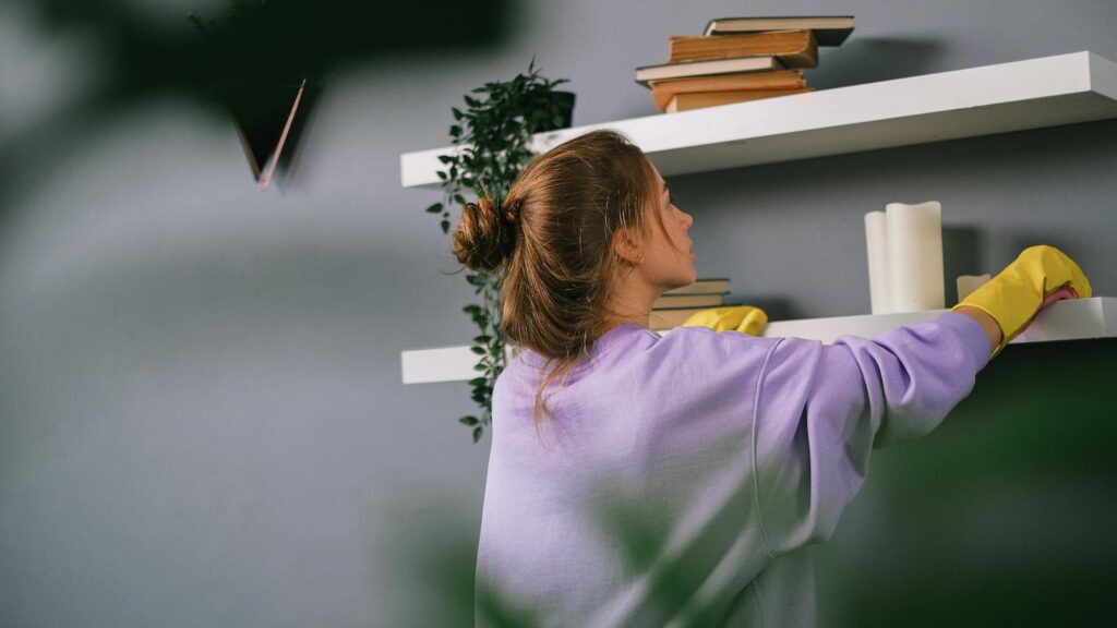 Woman in purple sweater dusts a bookshelf with candles and books.