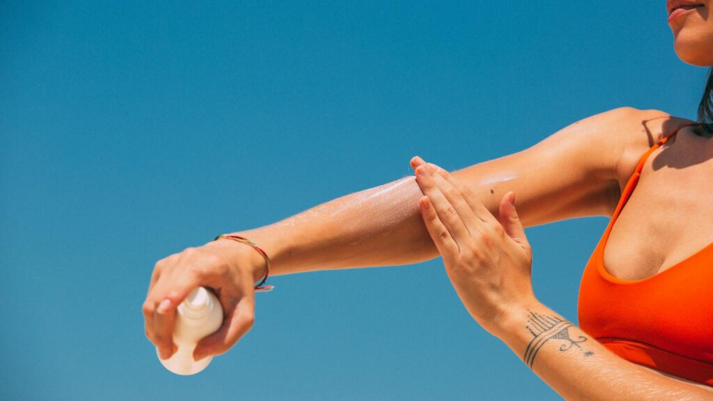 Close-up of a woman applying sunscreen on her arm under a clear blue sky at the beach.