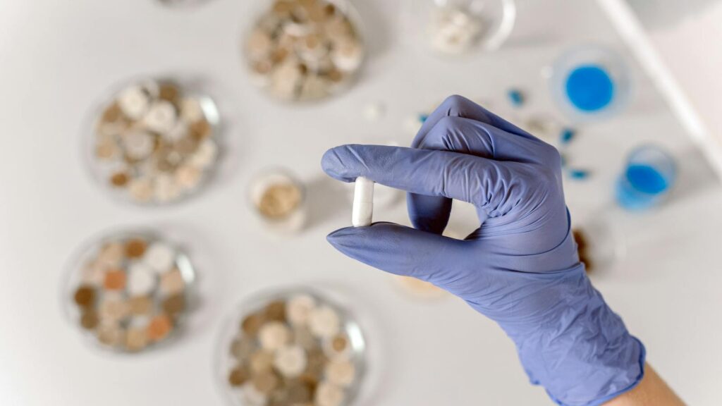 Close-up of a gloved hand holding a white capsule in a lab environment.