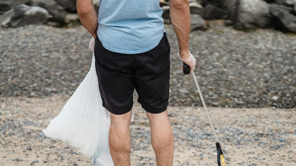 A man helps the environment by picking up plastic litter on a rocky beach with a grabber tool.