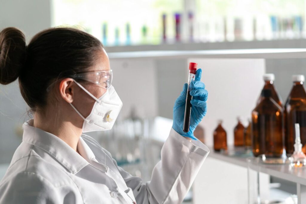 A female scientist wearing protective gear examines a vial in a well-lit modern laboratory.